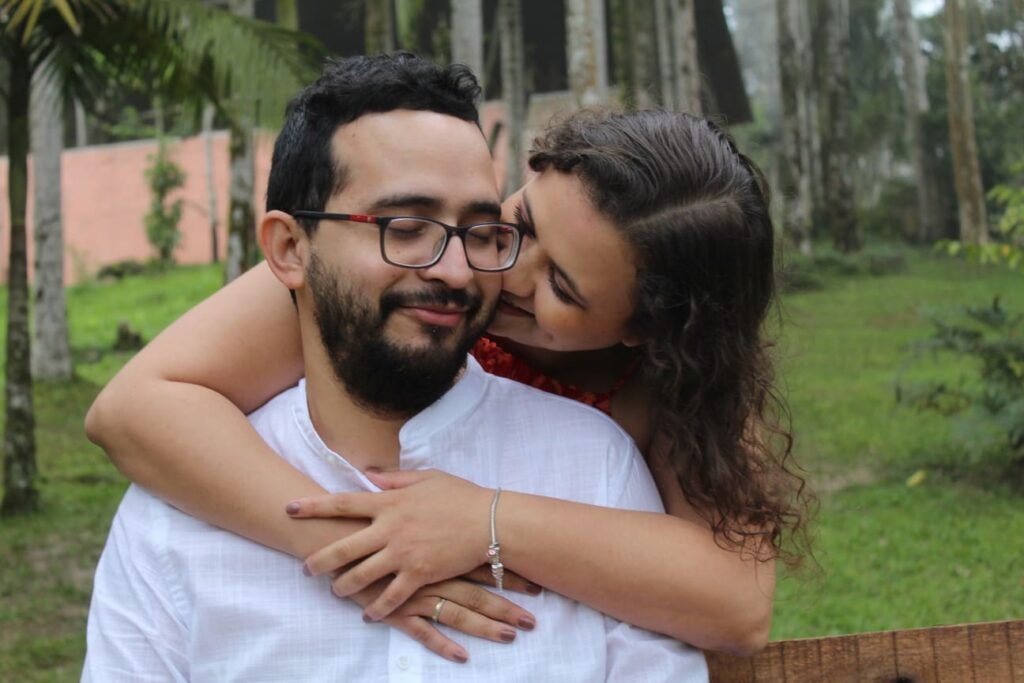 Casal sorridente em um momento de carinho ao ar livre. Uma mulher abraça o homem por trás, encostando o rosto em seu ombro, enquanto ele sorri de olhos fechados, usando óculos e camisa branca. Ao fundo, árvores altas e vegetação verde compõem um cenário natural e tranquilo. Ideal para transmitir amor, conexão e momentos felizes na natureza em um ensaio fotográfico de casal.