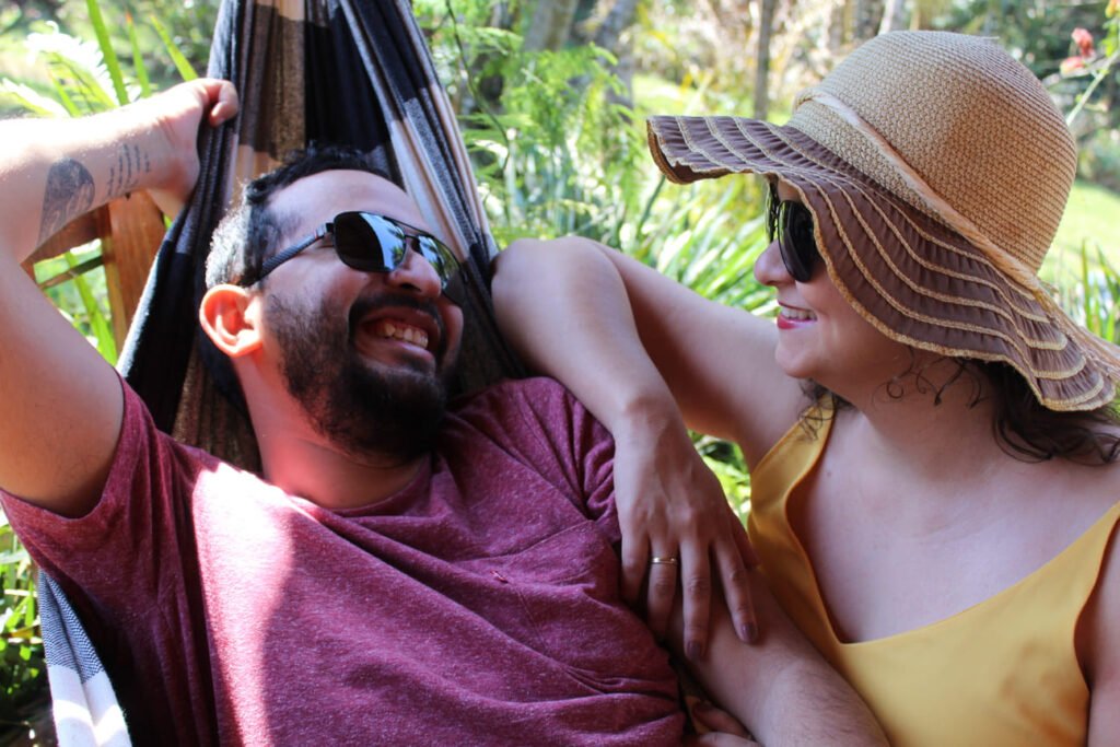 Ensaio fotográfico de casal relaxando em uma rede ao ar livre. O homem, vestindo camiseta vermelha e óculos de sol, sorri enquanto olha para a mulher, que usa vestido amarelo, chapéu de palha e óculos escuros. Eles estão rodeados por vegetação, em um clima descontraído e ensolarado. Ideal para temas de lazer, romance e dias de descanso na natureza.