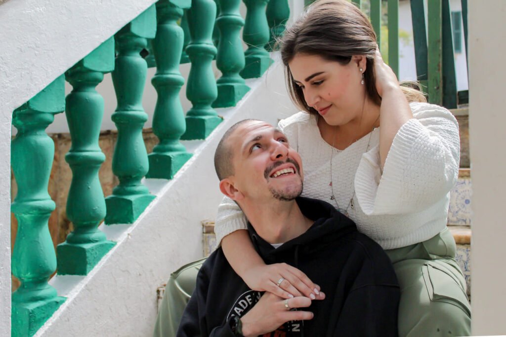 Casal sentado em uma escada branca com corrimãos verdes. A mulher, vestindo blusa branca, olha carinhosamente para o homem, que está sentado e a encara com um sorriso. Ele veste moletom preto e calça casual. A cena transmite romance e cumplicidade, perfeita para conteúdos sobre amor, relacionamentos ou sessões fotográficas urbanas.