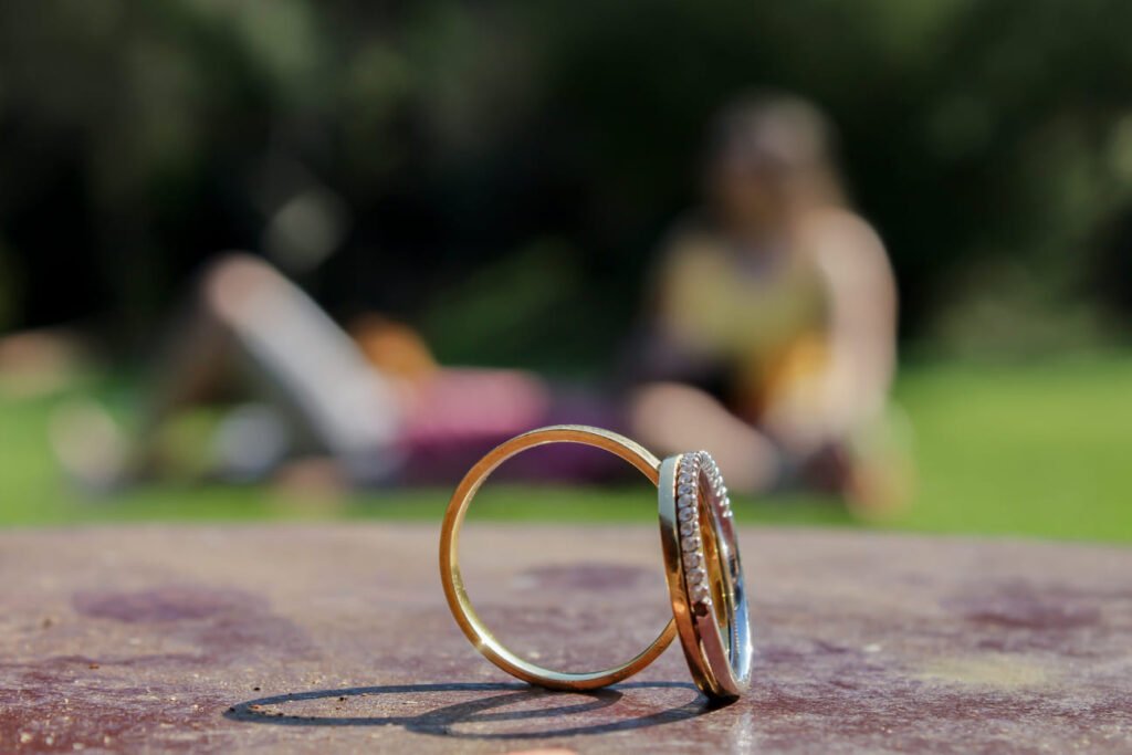 Fotografia de um anel de noivado em destaque, com fundo desfocado de natureza com um casal. Ensaio fotográfico pré-wedding celebrando o compromisso e o amor.