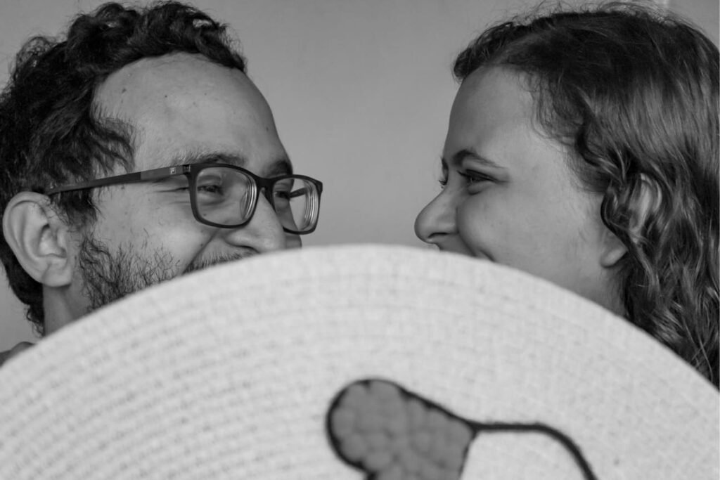 Casal sorridente compartilhando um momento íntimo enquanto seguram um chapéu de palha juntos, com fundo de natureza desfocado. Ensaio fotográfico pré-wedding com atmosfera romântica e leve.