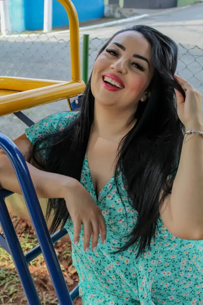 Fotografia de uma mulher jovem ao lado de um escorregador em um parque, sorrindo espontaneamente, com luz natural destacando o cenário descontraído. Ensaio fotográfico casual ao ar livre, capturando momentos autênticos e alegres.