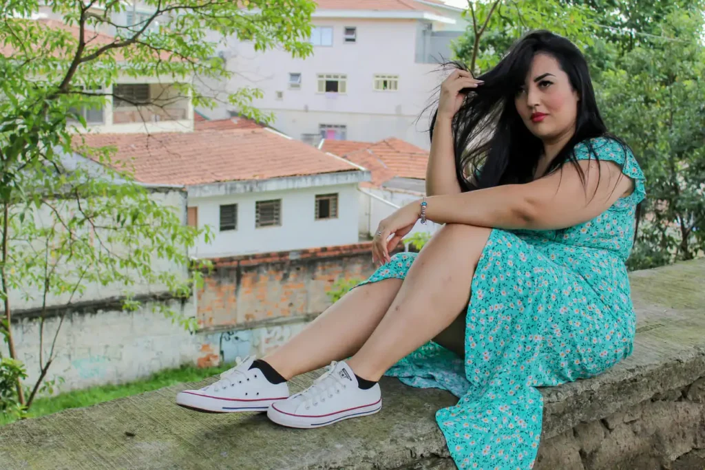 Mulher sentada em um ambiente externo, vestindo um vestido azul claro e tênis brancos, olhando para a câmera enquanto ajeita o cabelo em um ensaio casual.